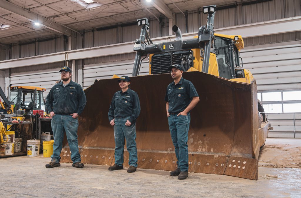 Three Diesel technicians proudly standing in front of a John Deere bull dozer